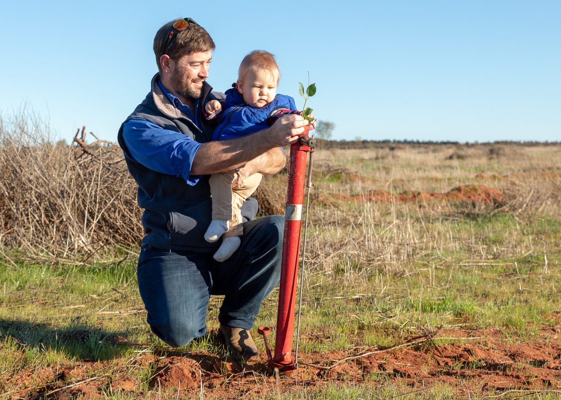 Man crouching holding a child in one arm and seedling at the top of a pottiputki planting tool in the other.