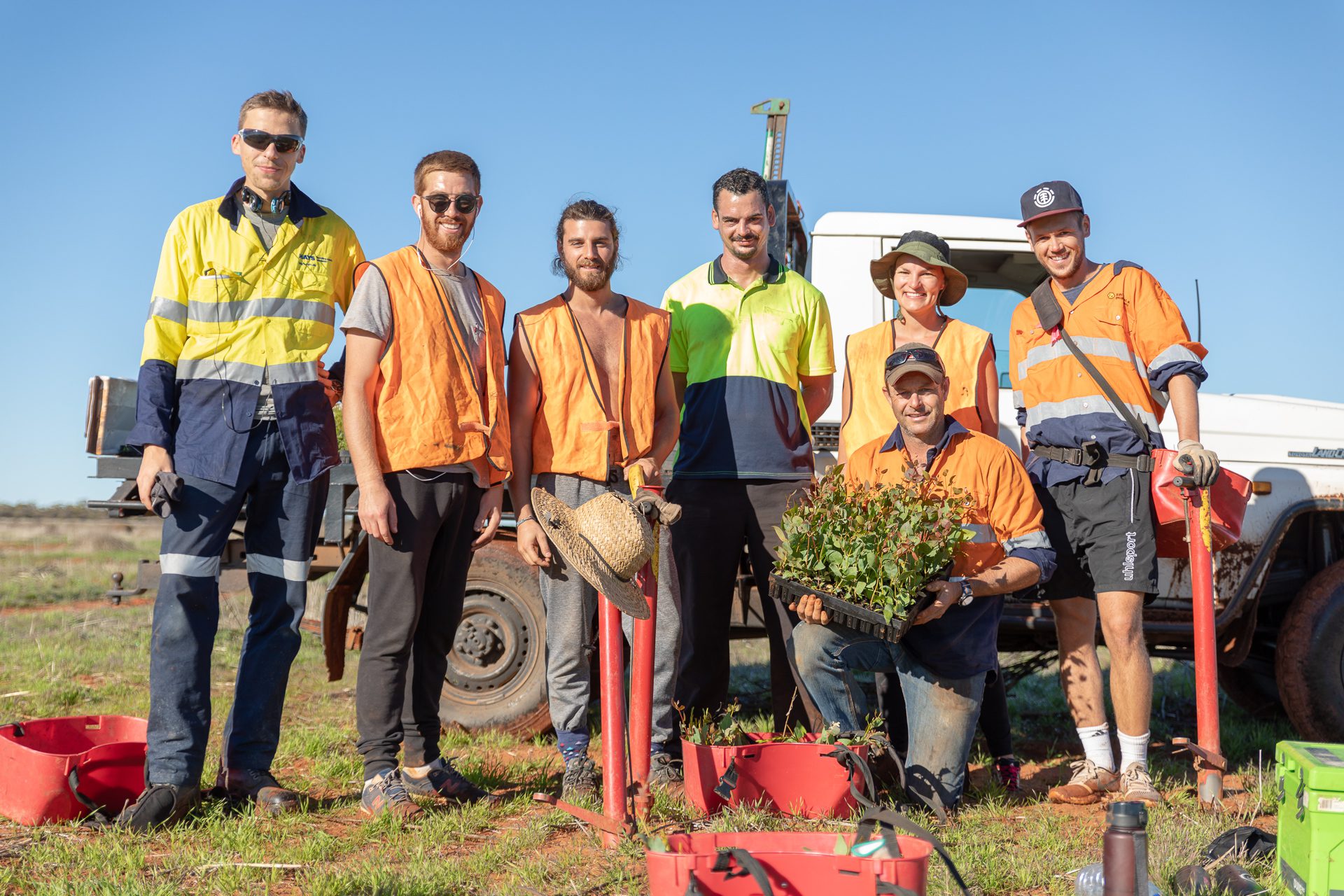 Seven people in high-vis clothing standing in front of a ute, with seedlings and planting tools.