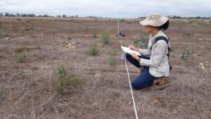 Woman kneeling in planting area writing on a piece on paper