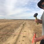 Man wearing glasses and a hat hammering a wooden stake into the ground of a sandy paddock.