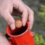 Close up photo of a persons hands holding a small round nut and a red metal tube.