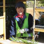 Lady sitting in a trailer wearing a navy blue beanie and high vis yellow vest holding a seedling