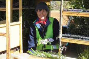 Lady sitting in a trailer wearing a navy blue beanie and high vis yellow vest holding a seedling