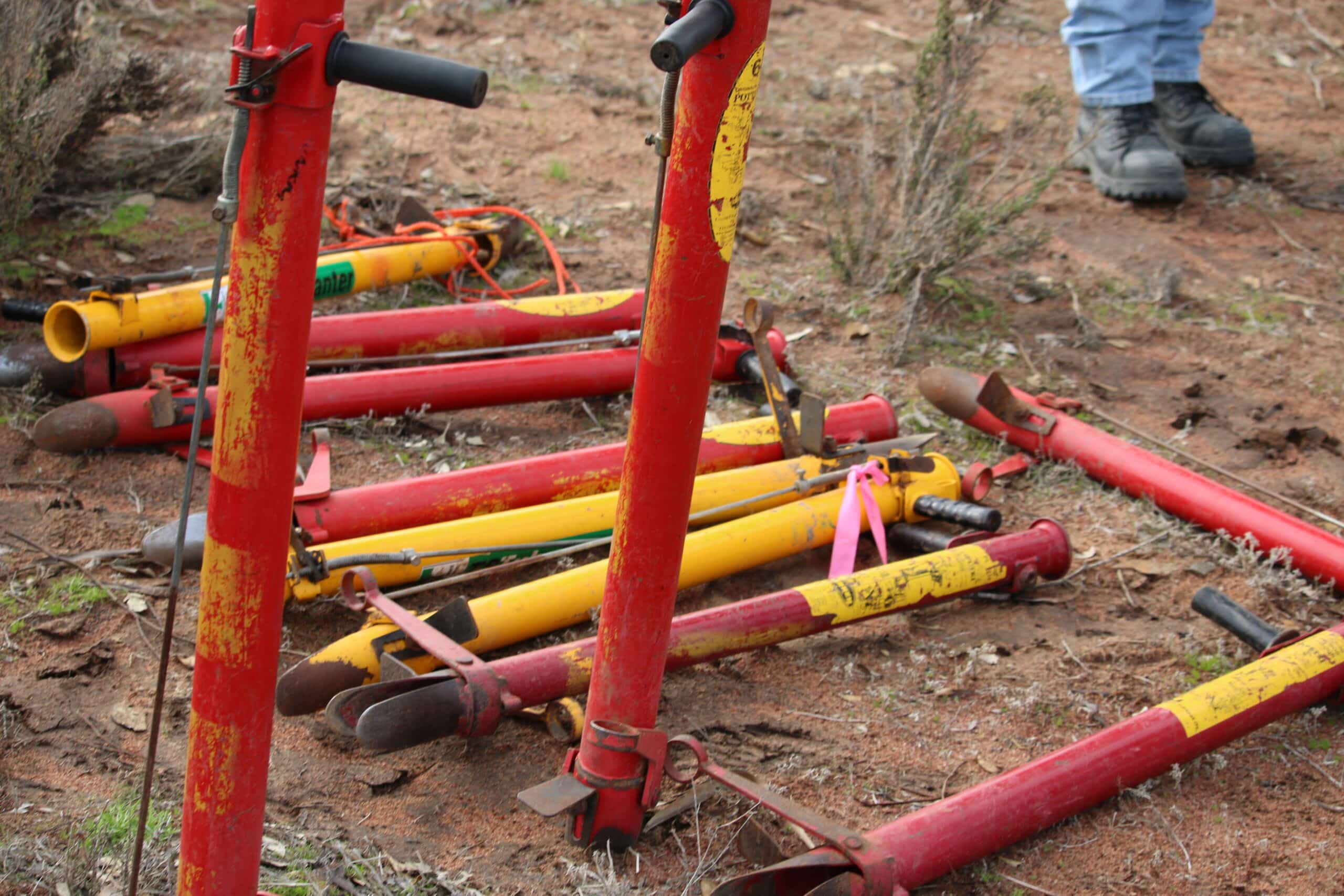 Two red coloured manual tree planting tools standing in the ground with several more lying horizontal in the background.