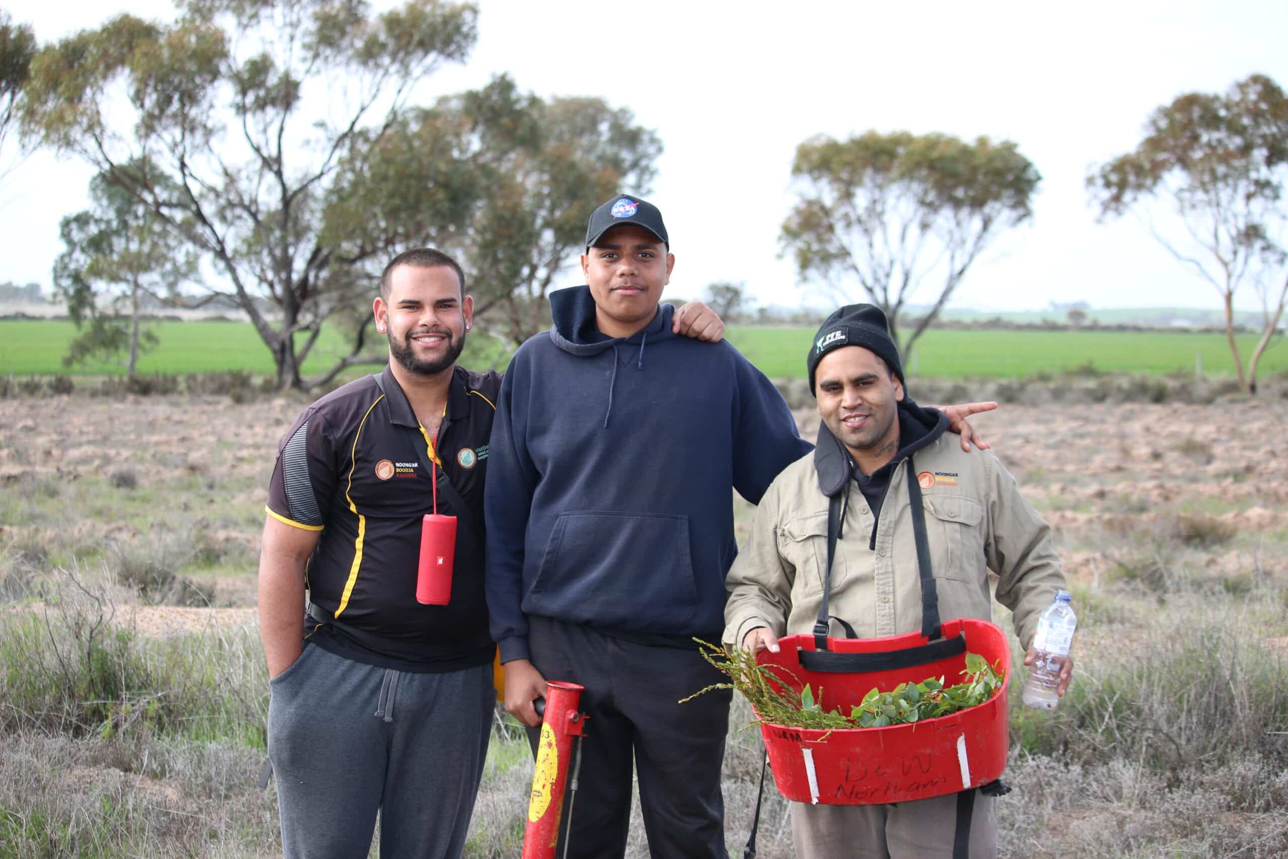 Three young men standing in a field. The man on the right is holding a red bucket full of seedlings.