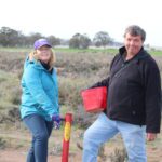 Man and a woman standing in a field during tree planting event. Woman wearing a bright blue jacket holding a pottiputki (hand-held tree planting device). Man wearing a black sweater holding a bucket full of seedlings.