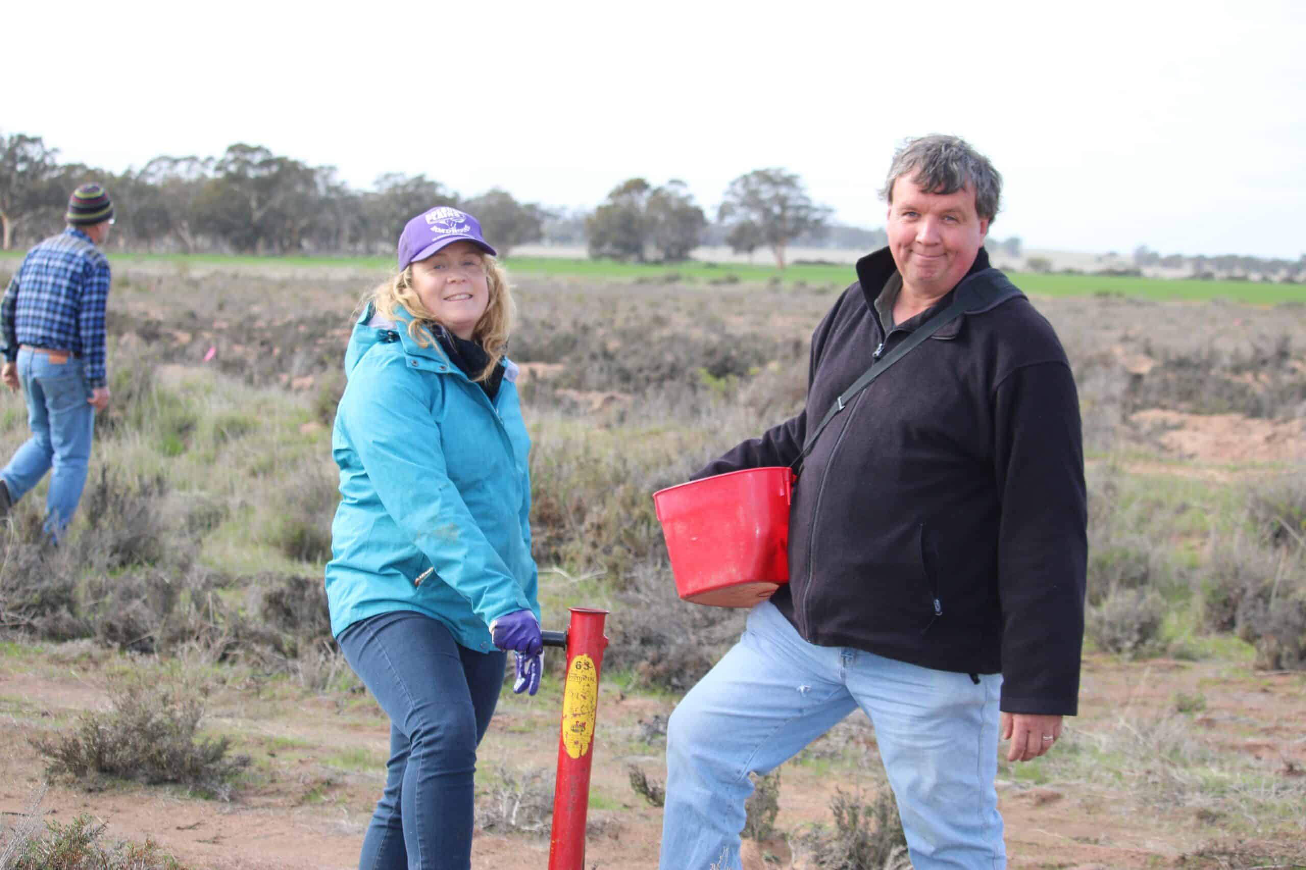 Man and a woman standing in a field during tree planting event. Woman wearing a bright blue jacket holding a pottiputki (hand-held tree planting device). Man wearing a black sweater holding a bucket full of seedlings.