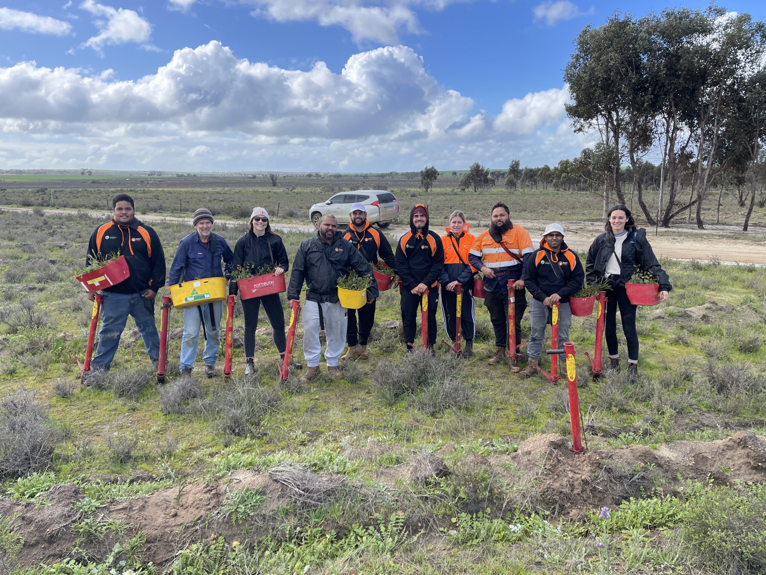 Group of ten people standing in a paddock holding buckets full of native seedlings and pottiputki planting devices