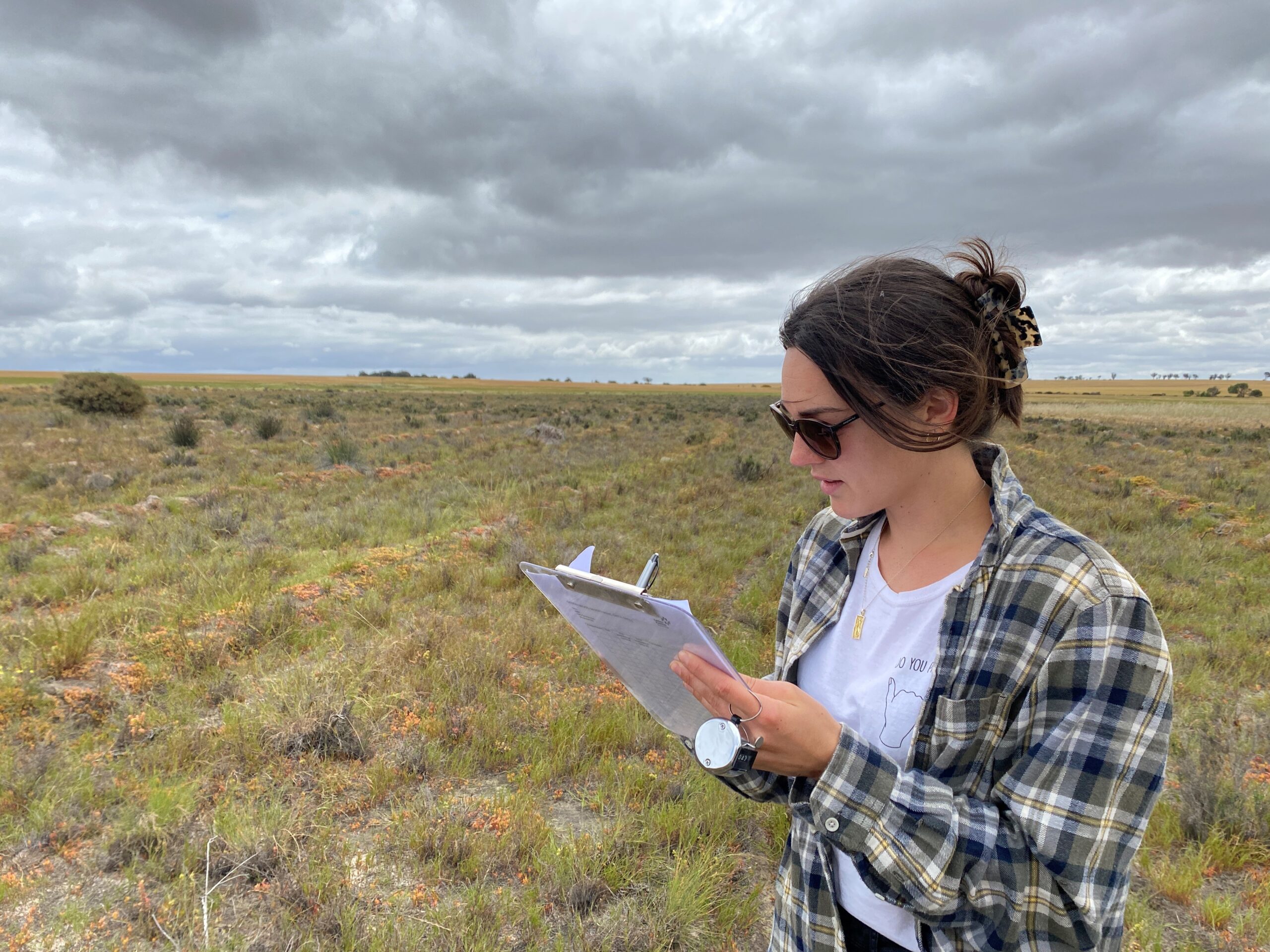 Girl wearing a white tee and flannel shirt standing in a paddock holding a clipboard and taking notes
