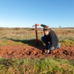 Woman crouching on red dirt next to a seedling holding a red pole