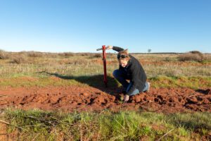 Woman crouching on red dirt next to a seedling holding a red pole