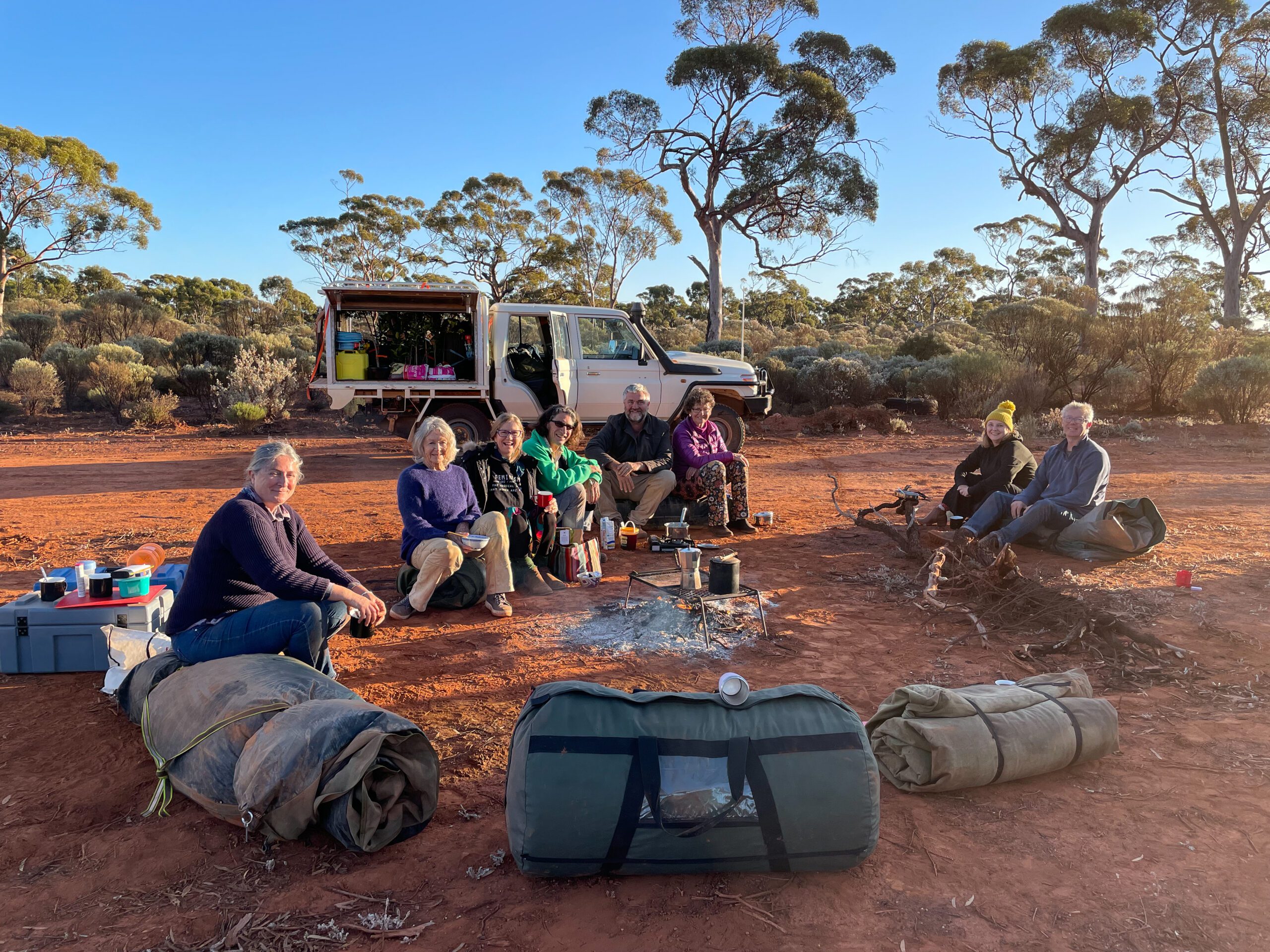 Eight people sitting around a fire in front of a white car on red dirt