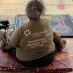 Indigenous woman sitting facing away from camera wearing green shirt with text "Carbon Positive Australia - Your Footprint, Australia's Trees, Our Future" on the back. Traditional dot art painting on ground in front of woman with paint supplies to the left.
