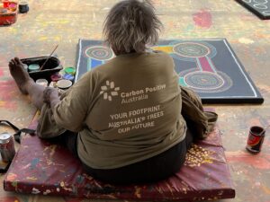 Indigenous woman sitting facing away from camera wearing green shirt with text "Carbon Positive Australia - Your Footprint, Australia's Trees, Our Future" on the back. Traditional dot art painting on ground in front of woman with paint supplies to the left.