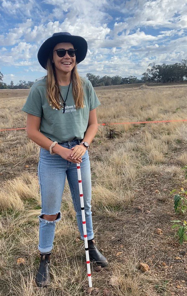 Young woman standing in a field holding a measuring stick to conduct monitoring at a carbon planting site.