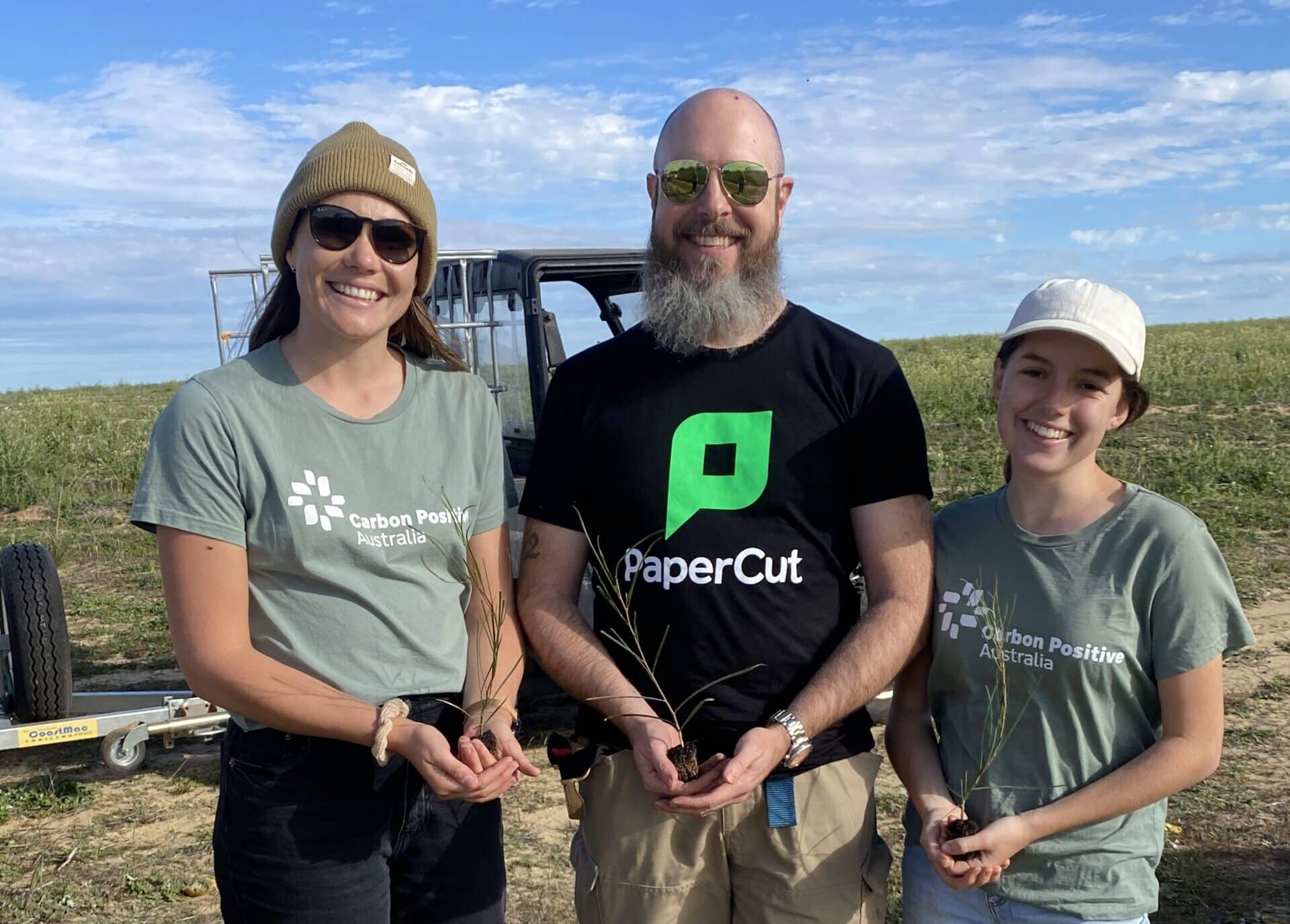 A man wearing a black t-shirt with "PaperCut" written on the front flagged by two women wearing green shirts with "Carbon Positive Australia" on the front. All three are holding a seedling out to the camera.