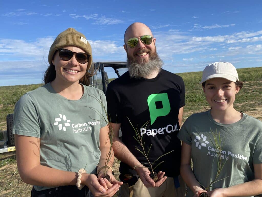 A man wearing a black t-shirt with "PaperCut" written on the front flagged by two women wearing green shirts with "Carbon Positive Australia" on the front. All three are holding a seedling out to the camera.