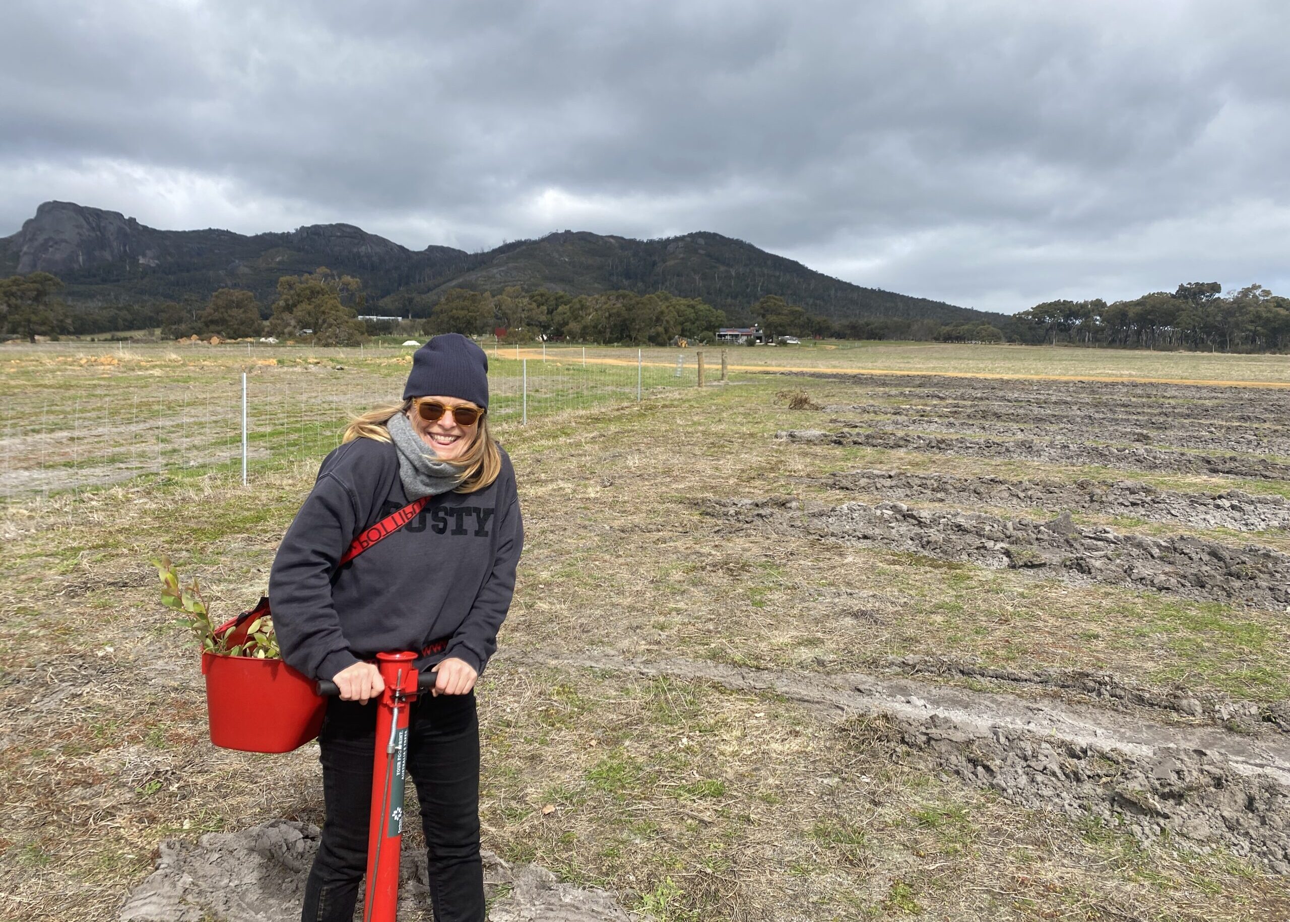Lady holding a tree planting device and a bucket of seedlings with mountain range in the background