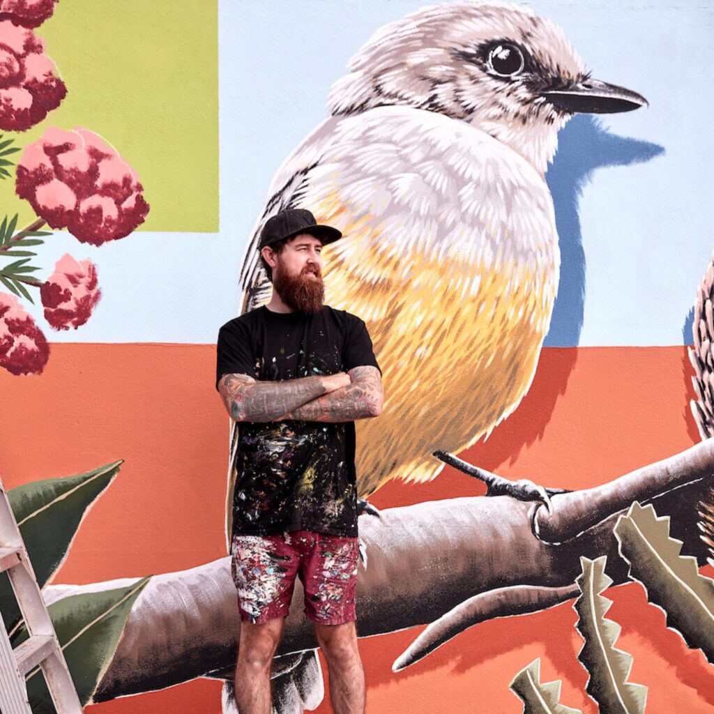 Perth-based muralist Brenton See in front of a mural of an Australian native bird and flowers.