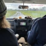 Two people sitting in the front seats of a car driving on a dirt road