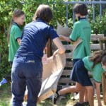 Women and kids holding cardboard outside with wood pallets in abckground and kid holding a watering can