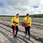 Two women in yellow RAC shirts standing in the middle of a planting row with a seedling bucket and pottiputki.